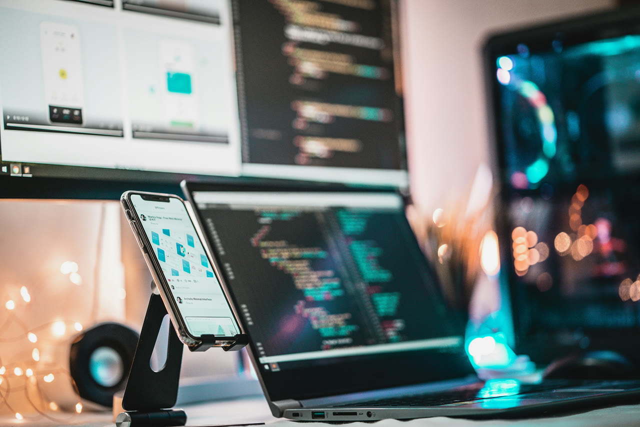 A modern desk setup with a smartphone displaying a UI design, a laptop showing code, and a large monitor in the background, symbolizing digital work and technology solutions.